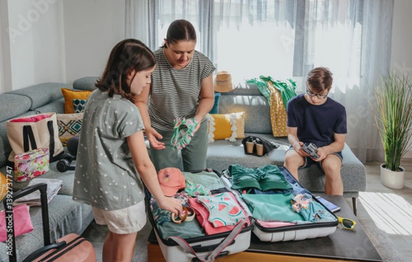 Fototapeta Family folding and packing clothes and beachwear into suitcases while teenage male using digital camera, getting ready for summer vacation. Mother and siblings preparing bags for trip in living room.