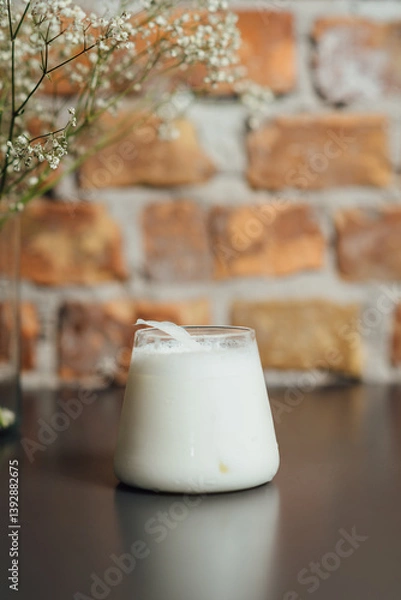 Fototapeta Creamy white drink served in a glass sitting on a dark surface, accompanied by delicate botanicals, emphasized by a warm brick backdrop, vertical composition