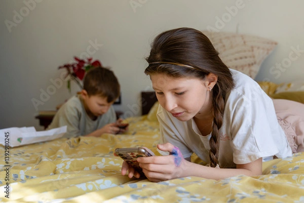 Fototapeta Kids using smartphones while lying on bed. Young girl and boy lying on a bed, focused on their smartphones, suggesting screen time or digital entertainment at home.
