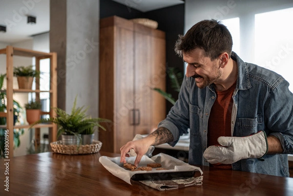 Fototapeta Happy man taking freshly baked cookies from baking tray in kitchen