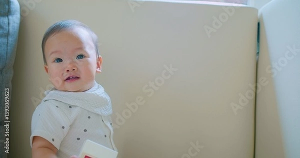 Obraz Cute baby with a curious expression holding a toy wearing a white outfit and scarf sitting indoors.