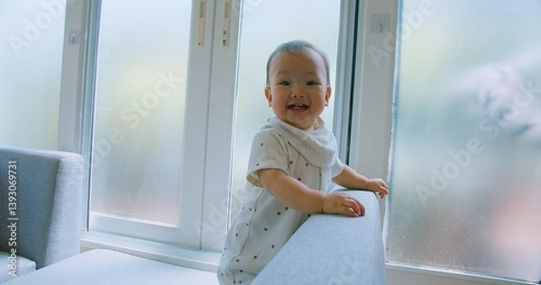 Obraz Curious Asian Baby Exploring Home Environment on Sofa in Bright Living Room with Natural Light with a curious expression.