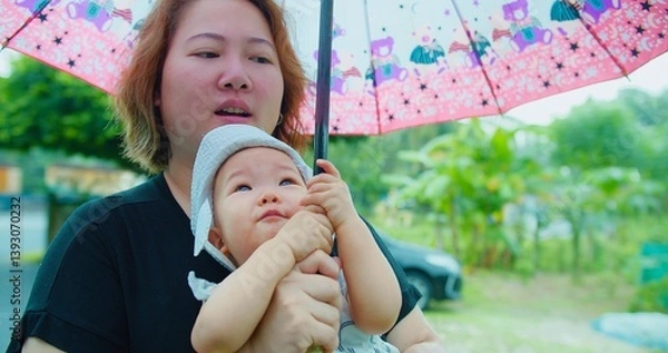 Obraz A Asian mother and her baby Enjoying share a moment under a vibrant umbrella on a rainy day, surrounded by lush greenery.
