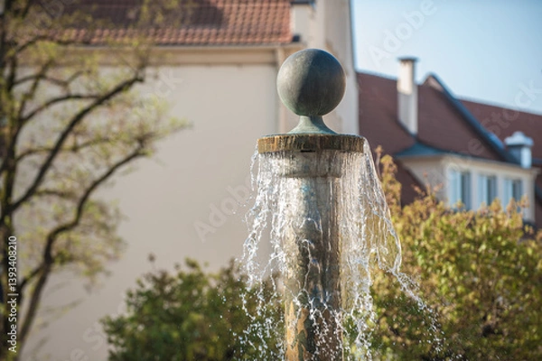 Obraz Brunnensäule mit Kugelabschluss und sprudelndem Wasser vor Wohngebäude mit Laubbäumen mit Herbstlaub in Nürtingen