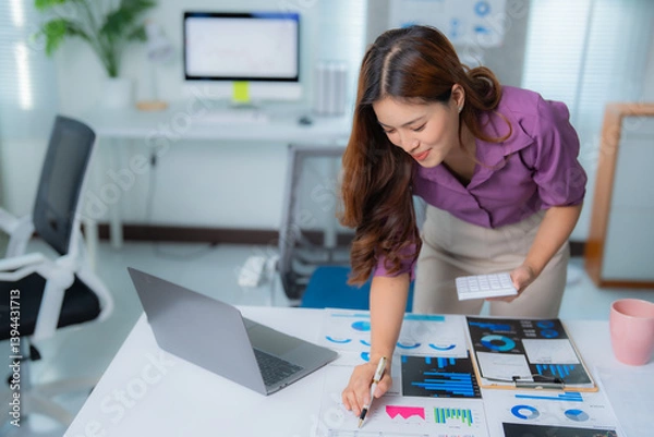 Fototapeta Young asian businesswoman holding calculator in hand and pointing at charts while analyzing marketing data in modern office with laptop and desktop computer