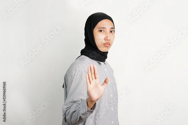 Fototapeta Young Asian woman raising her hand with palm facing forward in a stop gesture, expressing refusal or warning with a serious expression while standing against a plain background.