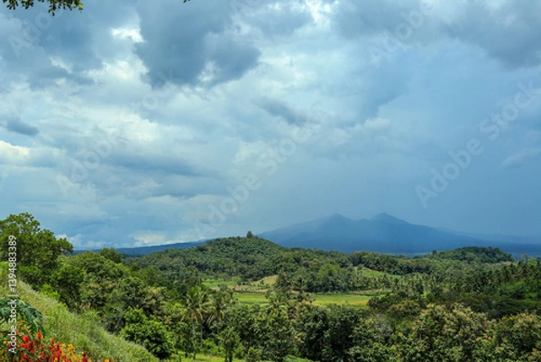 Obraz A wide-angle shot of a lush green forest valley stretching to a distant mountain range, with a dramatic sky of dark, stormy clouds overhead.