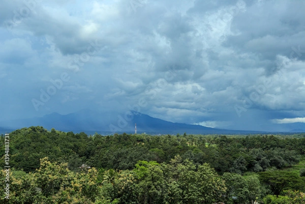 Obraz A wide-angle shot of a lush green forest valley stretching to a distant mountain range, with a dramatic sky of dark, stormy clouds overhead.