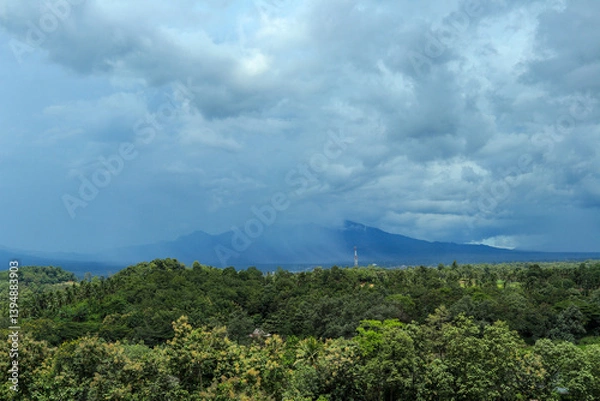 Obraz A wide-angle shot of a lush green forest valley stretching to a distant mountain range, with a dramatic sky of dark, stormy clouds overhead.