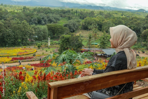 Obraz A woman wearing a beige hijab sits on a wooden bench, gazing out at a colorful flower garden.