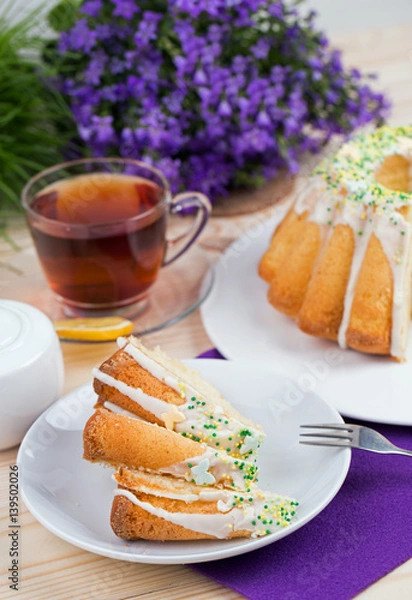 Fototapeta Homemade cake with icing sugar sprinkles on a plate and cup of tea on a wooden table with purple flowers in the background