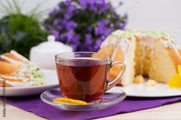 Fototapeta Homemade cake with icing sugar sprinkles on a plate and cup of tea on a wooden table with purple flowers in the background
