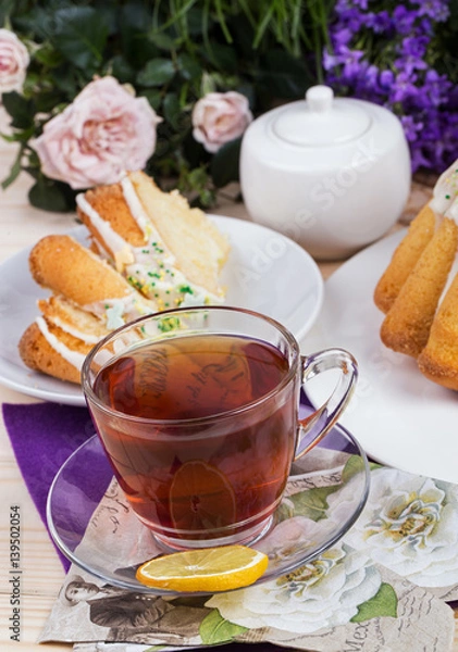 Obraz Homemade cake with icing sugar sprinkles on a plate and cup of tea on a wooden table with purple and rose flowers in the background