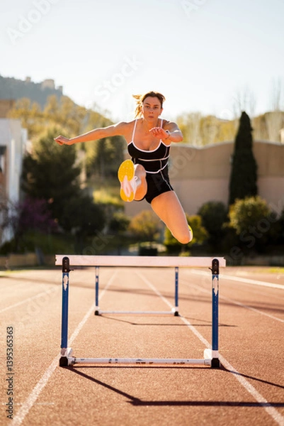 Fototapeta A Caucasian girl in her 20s and 30s is training on a running track.The girl is jumping a fence.Concept of female athletes.