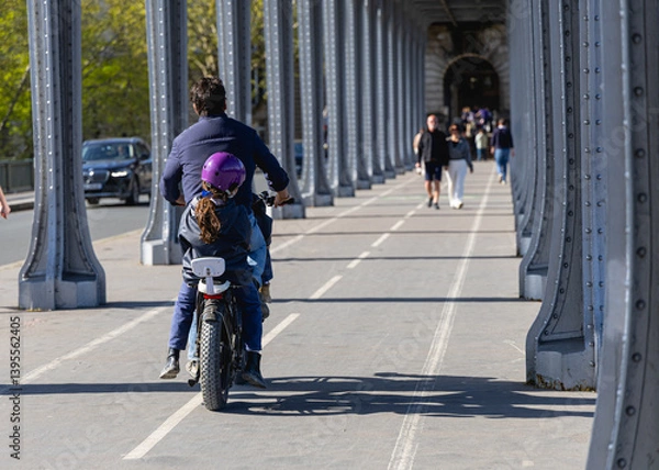 Obraz Un papa et sa fille en train de faire du vélo à Paris, sous le pont de bir hakeim, sur une piste cyclable. Le format de l'image est horizontal.