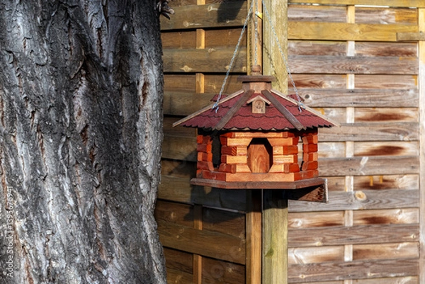 Fototapeta Wooden bird feeder made of wood in the form of a house hanging on a fence next to a tree.