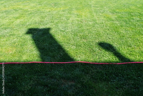 Obraz Shadow of a house on a well-maintained green lawn with a red rope laying on the grass. A conceptual image suitable for home security, real estate, outdoor safety, landscaping, and DIY content. 