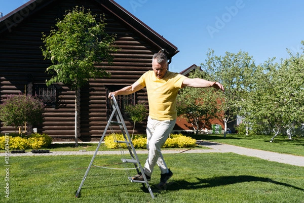 Obraz Senior man carefully stepping off a metal garden ladder on a sunny day in front of a wooden house. 