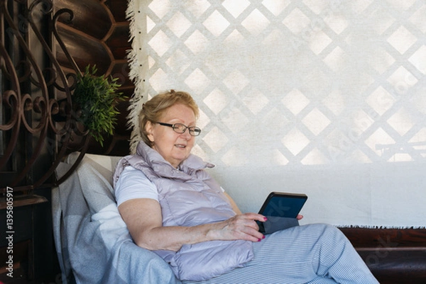 Obraz Senior caucasian woman relaxing on a cozy terrace, smiling while using a tablet or e-reader. 