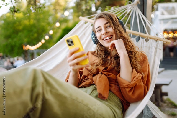 Fototapeta Beautiful woman relaxing in hammock by the lake wearing wireless headphones and using phone. Young woman enjoying sunny weather in park. Concept of relaxation, fun, blogging.