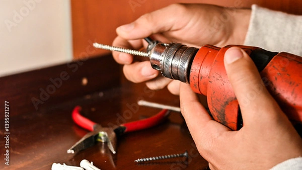 Obraz Hands holding a red power drill changing its bit on a wooden surface with other construction tools, emphasizing construction, DIY, and repair activities.