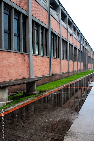 Obraz A contemporary brick building with rows of windows is reflected in water puddles after the rain, creating a balanced urban scene