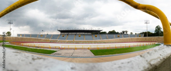 Obraz Empty cycling velodrome with yellow guardrails, set under a cloudy sky, surrounded by green nature.
