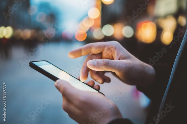 Fototapeta Close-up image of male hands using smartphone at night on city shopping street, searching or social networks concept, hipster man typing an sms message to his friends