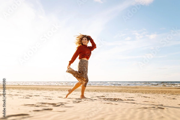 Fototapeta A beautiful woman with a hat walks on the beach at sunset. A young woman enjoys the seascape and feels free. The concept of relaxation, enjoyment.