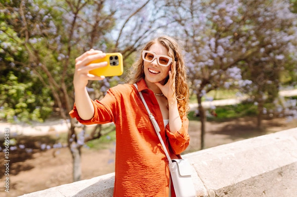 Fototapeta Selfie time. Beautiful woman taking photo or blogging on her phone on sunny day. Happy tourist enjoying vacation. Blogging, vacation concept.