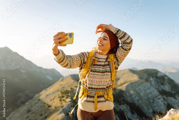 Fototapeta Hiker with bright hiking gear taking selfie on mountain. Cheerful woman feeling freedom and enjoying sunset on trail. Hiking, adventure concept. Active lifestyle.