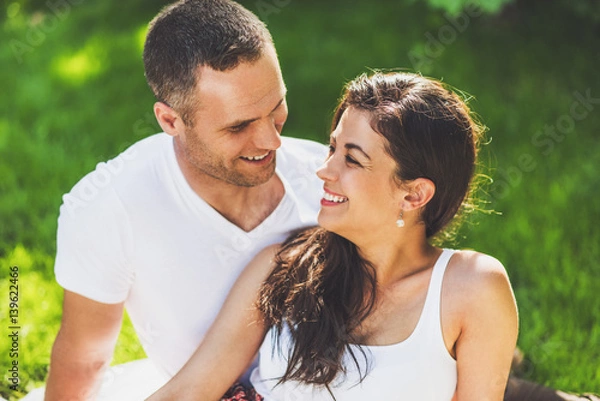 Fototapeta Close up portrait of beautiful couple in love sitting on a picnic blanket outdoor. Caucasian happy dreamy man and smiling brunette woman sitting on green grass in park looking to each other..