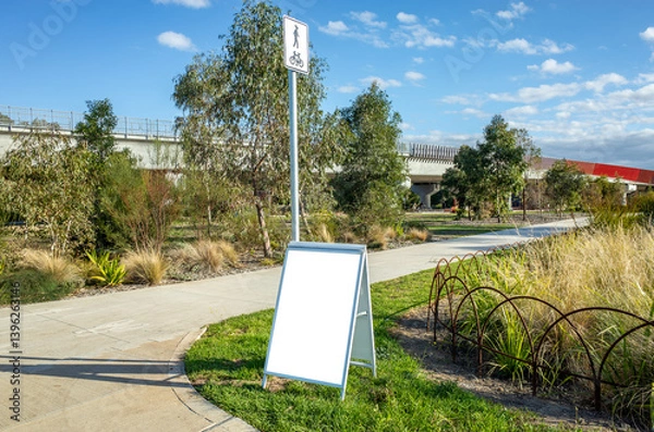 Obraz A-frame white directional signage standing alongside a biking and walking path in a public Australian park. Background texture of a blank mockup sign offers space for customizable messaging.