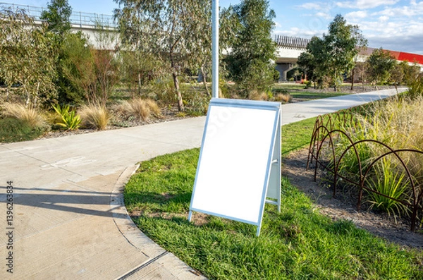 Fototapeta A-frame white directional signage standing alongside a biking and walking path in a public Australian park. Background texture of a blank mockup sign offers space for customizable messaging.