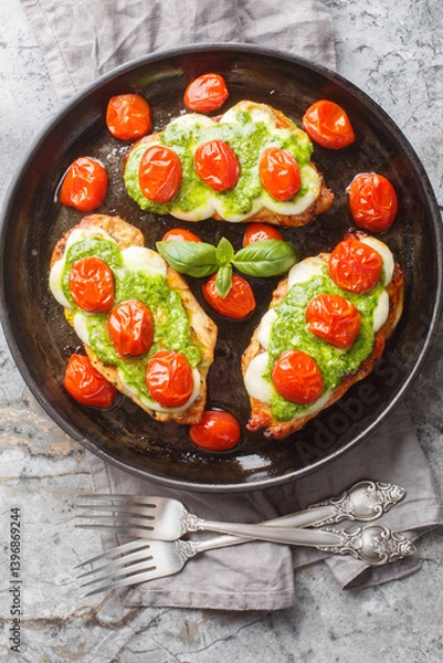 Fototapeta Margherita chicken that features pan-fried chicken breasts topped with melted mozzarella, pesto and tomatoes close-up on pan on table. Vertical top view from above