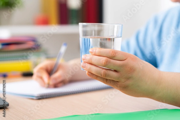 Fototapeta Close up of female student hand holding a glass of water studying with notebook at home