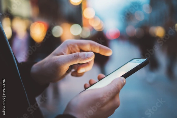 Obraz Zbliżenie męskich rąk wpisujących wiadomość SMS za pośrednictwem smartfona na zewnątrz, Hipster Man cieszący się wieczornym spacerem i używaniem telefonu komórkowego, bokeh światła