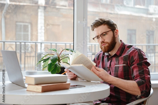 Fototapeta Concentrated bearded young man reading books while using laptop