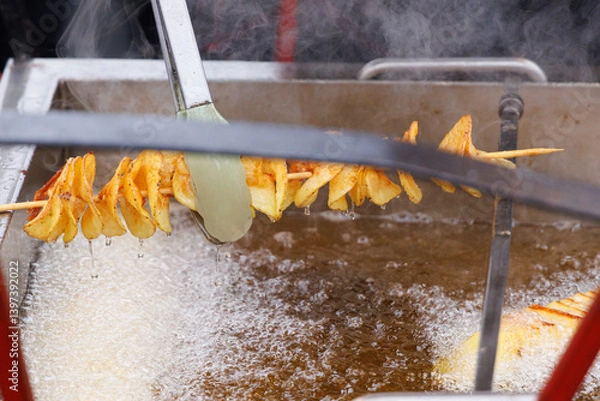 Fototapeta Fried potatoes in the form of a spiral on a wooden skewer are taken out of boiling vegetable oil.