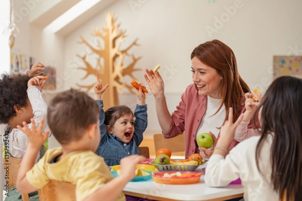 Fototapeta Smiling teacher and diverse group of preschoolers raise their hands and enjoy healthy snacks together, promoting fun, nutrition, and inclusion in a vibrant early learning environment.