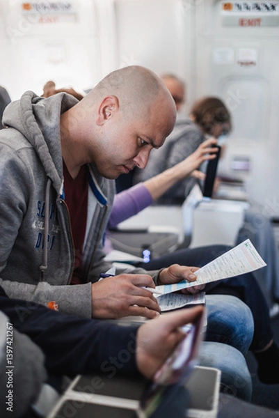Fototapeta Male traveler filling immigration form on flight to visit destination country sitting in airplane. Man is writing entry permit or visa on a paper on a flight. Passenger signing document on board.