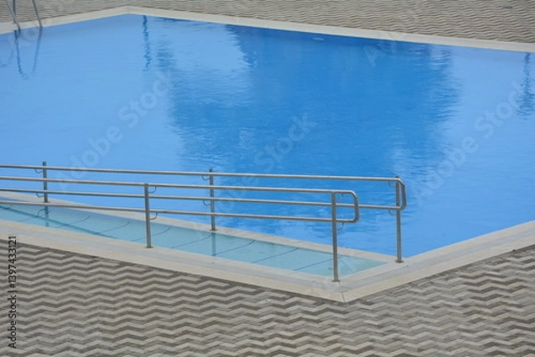 Fototapeta Poolside view with a smooth blue surface and metallic railing at a modern facility during daytime