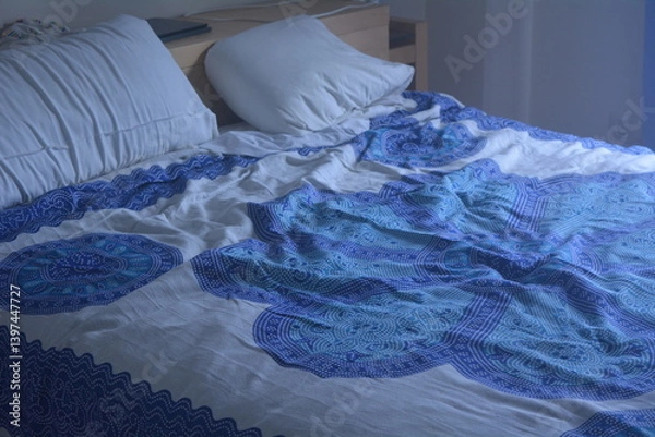 Fototapeta Cozy bedroom featuring a blue patterned bedspread with disheveled bedding in soft morning light