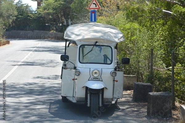 Fototapeta Classic white tuk-tuk parked on a quiet road surrounded by greenery on a sunny day