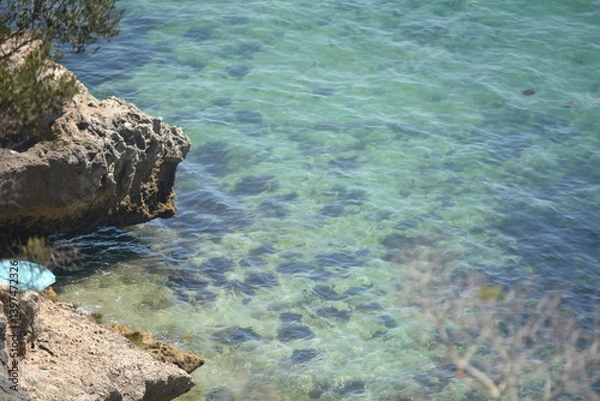 Fototapeta Clear turquoise water laps against rocky coastline under bright sunlight near a secluded beach in summer