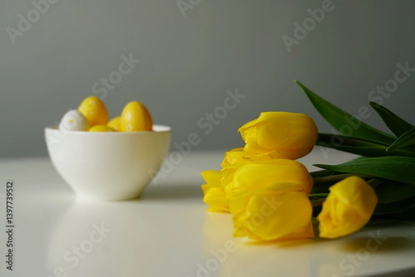 Obraz Bright Easter composition with yellow and white eggs in a white ceramic bowl next to fresh yellow tulips on a white table. Minimalist holiday still life, perfect for spring-themed content. 