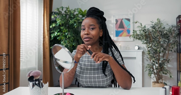 Obraz A makeup artist with dreadlocks paints her eyelashes while sitting at her desk. She records a vlog, sharing mascara benefits and beauty tips with her online audience.