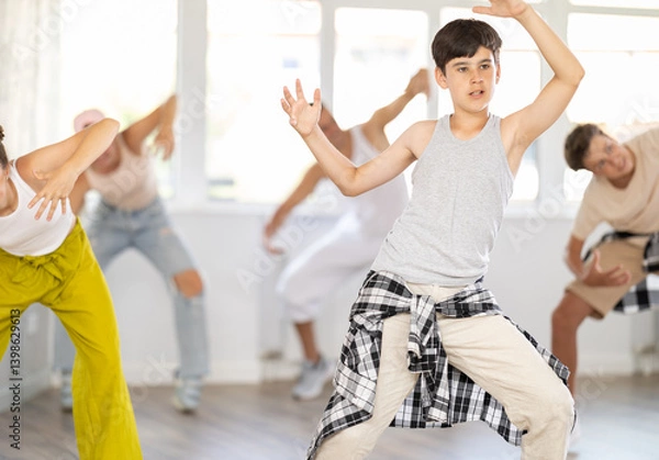 Fototapeta Happy teenage boy practicing basic hip-hop moves in training hall during group dancing classes