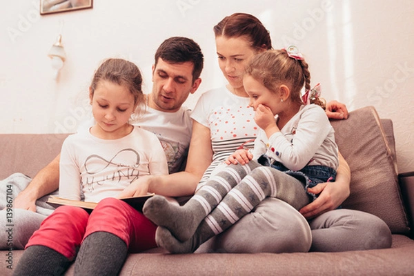 Fototapeta Mom dad and daughter read a book at home on the couch