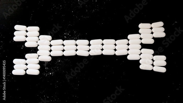 Fototapeta Oval-shaped white supplement tablets arranged in the shape of a bone. With black background.